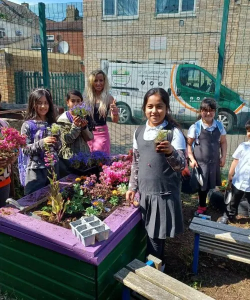 A group of children and an adult stood around a raised flower bed holding plants.