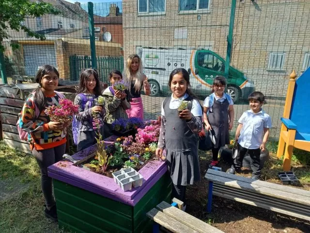 A group of children and an adult stood around a raised flower bed holding plants.