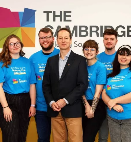 A man in a suit surrounded by people in blue shirts with The Cambridge Building Society's logo. They are standing in front of a white wall with The Cambridge's logo.