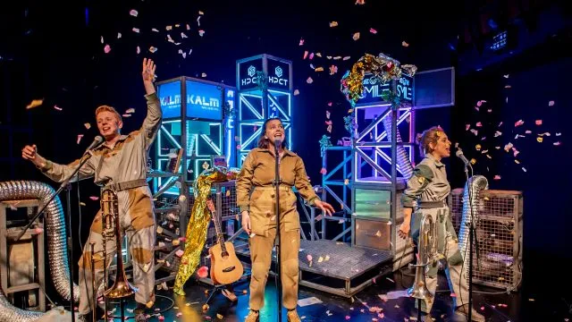 Three performers on stage signing against a dark blue background with LED lights and confetti in the air.