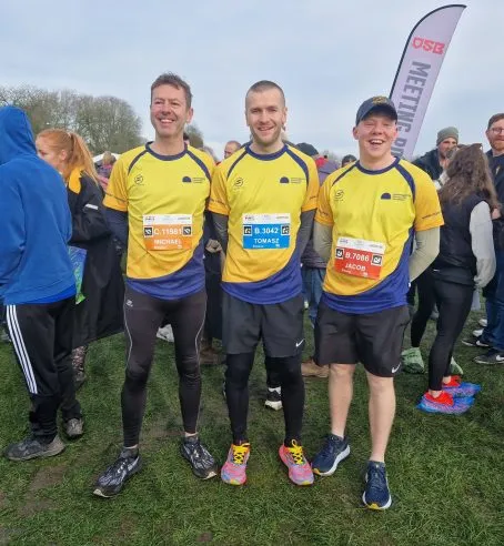 Three people standing side by side in running gear and Cambridgeshire Community Foundation branded yellow and blue tops. They're standing on grass with many people behind them.
