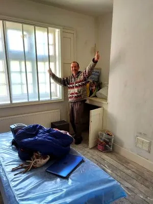 A person standing by the windows in a room, with his hands up in the air. His belongings are in