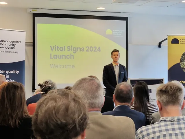 A room filled with people seated facing a speaker standing behind a podium with a presentation projected behind them and pop-up banners on both sides of the podium.